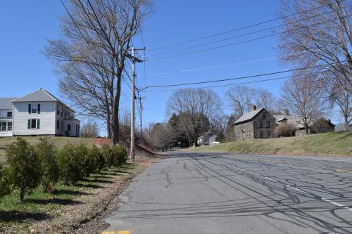 Same inn in the distance, now with a paved road. A neat row of trees has replaced the fence, with another house on the other side. Color photo.