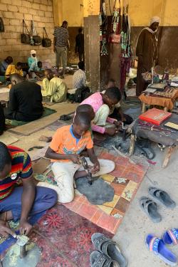 Four boys sit on the ground bent over leather mats. They are using hammers and chisels to shape small pieces of metal. In the room behind them older men work also on the ground and a few men stand to watch the artisans work.
