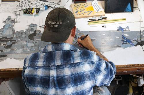 A man, facing away, hunches over a desk drawing an intricately detailed rendition of a gray Navy ship.