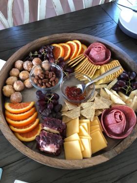 Close-up on a circular charcuterie board: orange slices, squres of cheese, bowls of nuts and marmalade, crackers, grapes, and slices of salami formed into the shapes of roses.