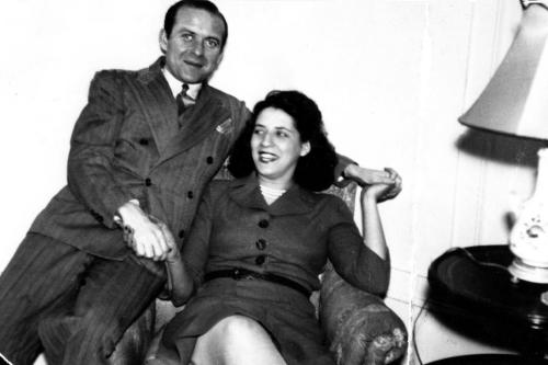A man, sitting on the arm of a chair, puts his arms around to pose with the woman in the chair. Black-and-white family photo.