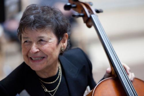 A woman wearing a black suit and gold necklace smiles and holds the neck of a cello.
