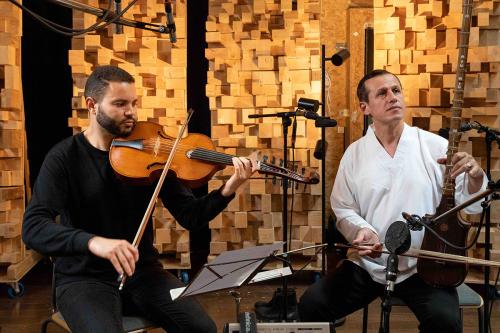 Two people perform in a recording studio, one on a large violin-type instrument, and the other on an upright, long-necked bowed string instrument.