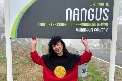 A woman with black hair and red, black, and yellow sweater poses under a roadside sign that reads Welcome to Nangus, Part of the Cootamundra-Gundagai Region, Wiradjuri Country.