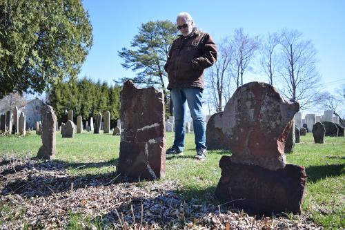 A man in jacket and dark sunglasses standing in a graveyard, looking down at one worn tombstone.