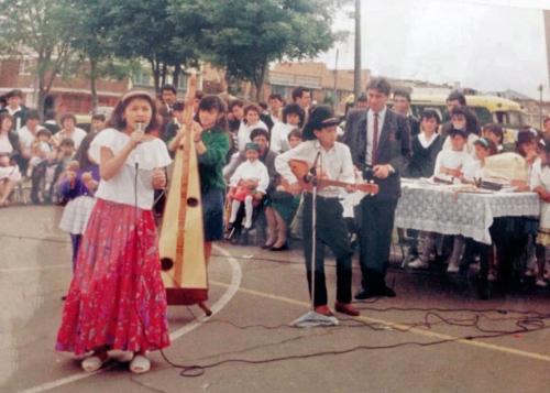 Child musicians perform outdoors on a blacktop, including a young girl singer in white blouse and long pink skirt. Behind her, kids playing maracas, harp, and cuatro, and a crowd of kids and adults.