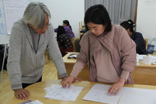 A man and a woman stand next to each other in front of a table. On the table are placed white sheets of paper with graphite sketches on them.