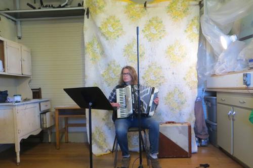 A woman with long dirty blonde hair, plaid shirt, and jeans plays a pearly white accordion, reading from a music stand. Behind her, she has rigged up a floral quilt to dampen the sound. To  her sides are cabinetry and a small lamp pointed at her.