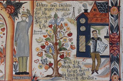 Detail from the painting, depicting Judy Larson. Script reads: Elders and children cut paper symbols for the Wycinanki Tree of Life. From the shed, Judy helps record and broadcast church service.
