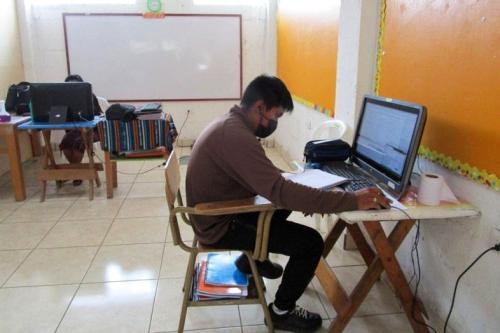 A young man sits at a desk in front of a computer with a notebook.