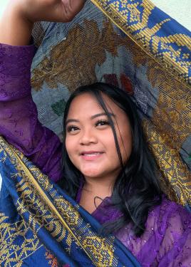A woman in a purple blouse holds up a translucent blue fabric embroidered with a gold pattern.