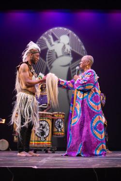 On a theater stage, a woman in a long purple dress hands a wisp of tan-colored horsehair to a man.