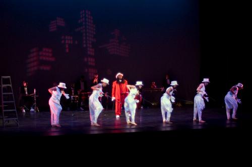 A dance performance on stage: six dancers dressed in all white in the front, with a person all in red right behind them. To the side of the stage is a ladder.
