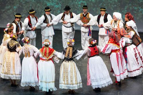 About 20 men and women, dress in traditional Croatian dance costume of white shirts, pants, and skirts, black vests and hats for the men and colorful scarves for the women, dance in a circle. Everyone holds hands with those two spots away, so their hands 
