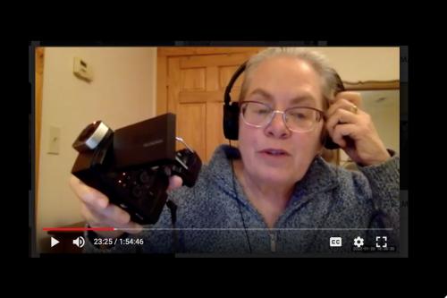 Screen shot of a video demonstrating how to use a video camera. In the center, a woman with grey hair and glasses holds up a small, black video camera to the screen.