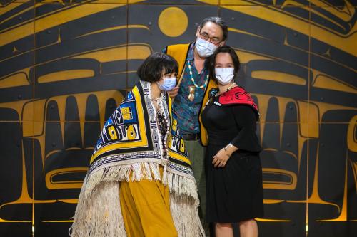 Parents and daughter embrace, all three wearing face masks and the daughter wearing ornate woven robe.
