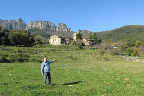 Man standing in an open green field, pointing toward the town and mountains behind him.