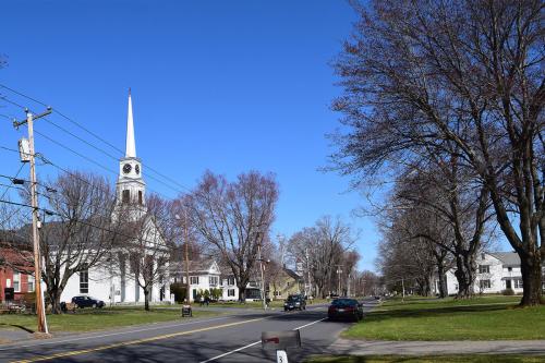 Same tree-lined street with cars instead. Church steeple is still visible. Color photo.