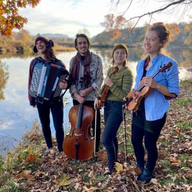 Four-piece band pose by the water under autumn leaves, holding their instruments: accordion, cello, viola, and violin.