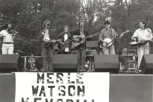 Six men on an outdoor stage playing fiddle, mandolin, banjo, and electric, acoustic, and steel guitar. A banner on the front of the stage reads Merle Watson Memorial.