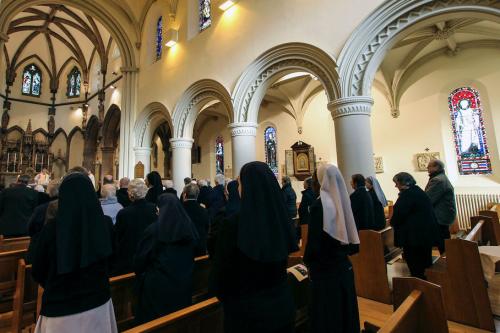 From behind, men and women standing up in pews of a church. Most are dressed in black, and a few of the women wear black or white veils.