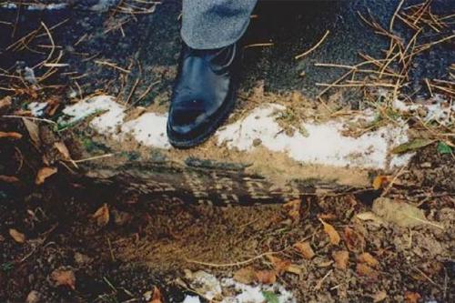 Closeup of someone stepping on the edge of a sidewalk, which is made from an old grave marker. It is obscured and covered in leaves and dirt.