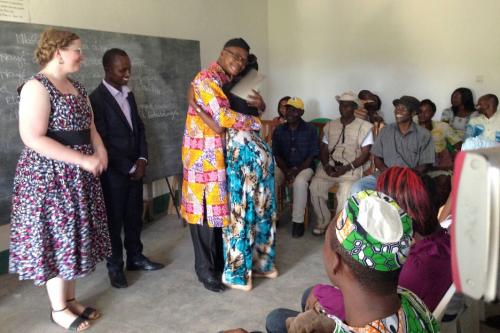 In the center of a classroom, a man wearing glasses, a black hat, and a yellow, purple, and red patterned tunic hugs a woman wearing a teal dress. In the man’s hand is a white piece of paper. They are surrounded by a smiling group of students and teachers