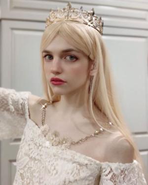 A person poses with a gold crown, long straight blond hair, gold necklace, and lacey white dress.