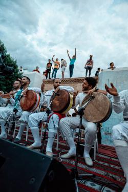 Men, in the midst of a performance, sit holding large hand drums. Behind them, youth stand on a roof dancing.