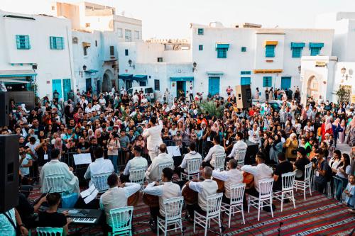 In a town square surrounded by white and blue buildings musicians have set up chairs to perform. Surrounding them is a large crowd of people, excitedly waiting for the show to begin.
