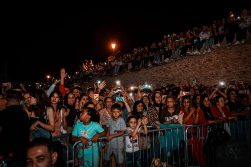 People of all ages stand pressed up against a barrier, watching a performance. They look engrossed and some hold cameras/cell phones to capture what’s happening.