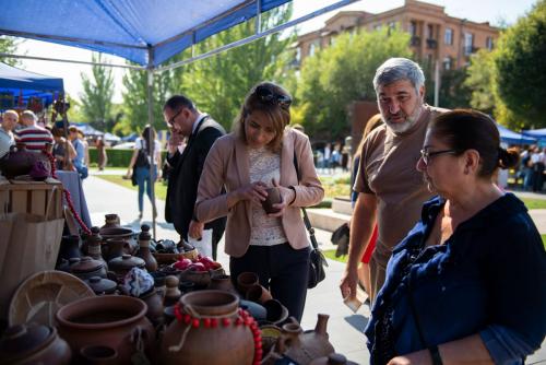 At an outdoor market, four people stand to the right of a table full of handmade pottery. A woman in the center of the image holds a small clay cup in her hands.