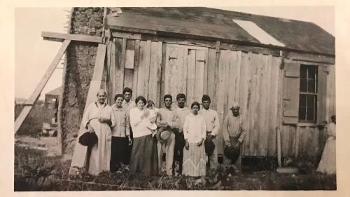 A large group of people, all in white, poses outside a single-story wooden building with stone chimney. Black-and-white photo.