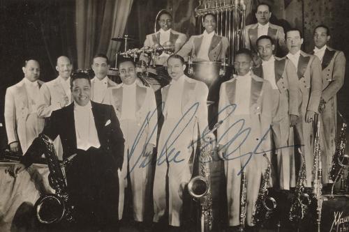Thirteen Black men pose on stage with instruments. Twelve are in light-colored suits, and Duke Ellington in front wears black. Black-and-white photo with Duke’s signature in blue over it.