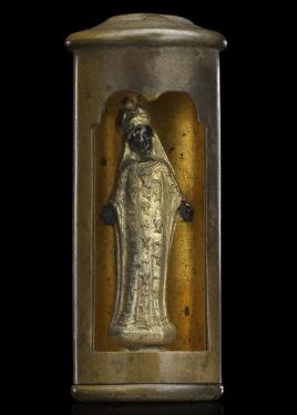 A trench art sculpture featuring a Black Madonna figurine with outstretched arms in the hollow interior of a bullet casing. A rectangle has been cut into the side of the casing with a scalloped edge on top. Inside, a Madonna figure stands in a long gold d