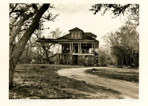 A black and white photoprint of a house that is still standing but has suffered severe damage from Hurricane Katrina.