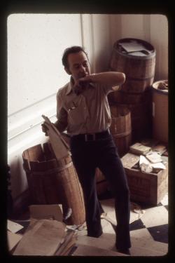 A man looks surprised in a room filled with barrels and loose paper, maybe a theatrical set.