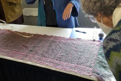 People inspect a pink and green woven shawl laid flat on a table.