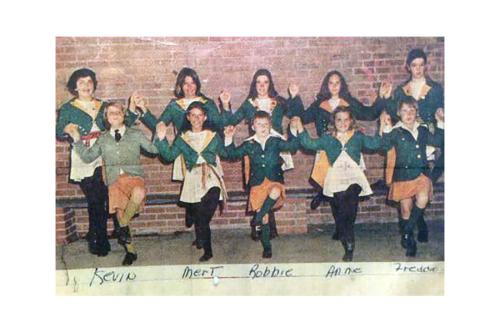 Old color photo of a dance class posing, mid-step. They are wearing matching green, white, and orange outfits. Some names are written at the bottom: Kevin, Mert, Robbie, Anne, Freddie.