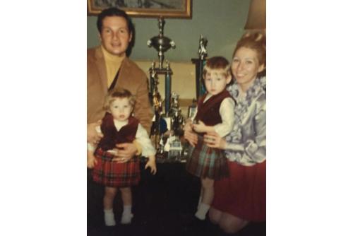 Old color photo of parents posing with two toddlers, both dressed in kilts and vests. Between them are several trophies. 