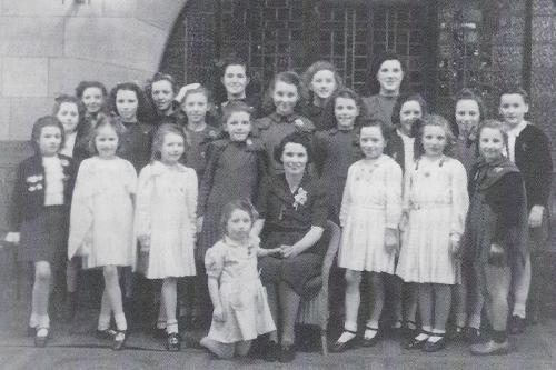Old black and white photo of young boys and girls posing with their teacher, seated center.