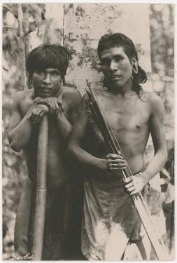 Two bare-chested men with thick dark hair and elongated pierced ears stand side by side. One leans on a long bamboo tube. The other holds a bunch of long wooden arrows. Black-and-white photo.