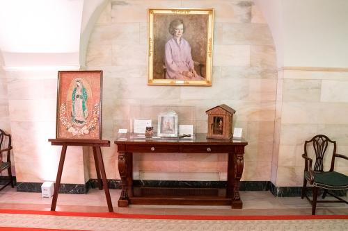 A display on a table and easel, set against a white marble wall and below a painted portrait of a woman. On the table are various small altar objects. To the left, on an easel, is a portrait of the Virgin of Guadalupe.