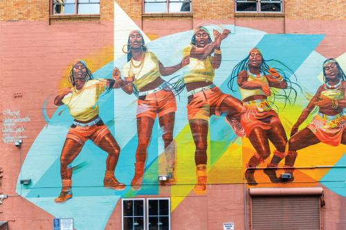 Mural painted on red brick wall, showing five version of the same Black woman, morphing from modern clothes T-shirt, jean shorts, sneakers, hoop earings, and headband) on the left to more traditional African looking dress (bandeau top, skirt adorned with 