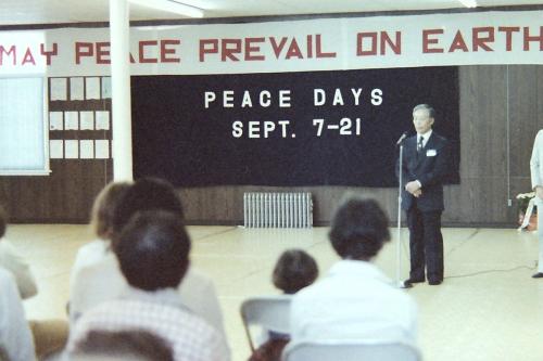 In an open studio room, a man in a black suit stands at a microphone, hands clasped in front of him, facing a seated crowd in the foreground. Behind him, banners read: May Peace Prevail on earth. Peace Days Sept. 7-21. Old color film photo.