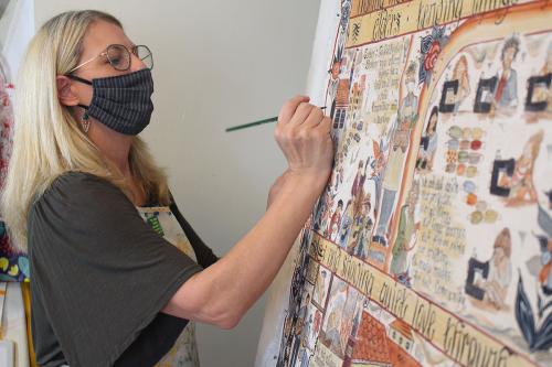 A woman with longe blond hair and a face mask paints a detailed scene on a large canvas.