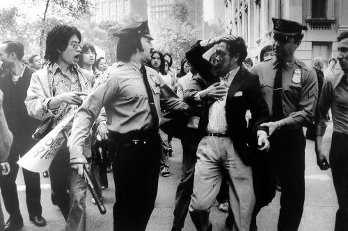 In the foreground, three police officers confront and hold back an Asian man who is holding his hair back, his face covered in blood. Behind them is a group of Asian men and women, one yelling at the police. Black-and-white photo.