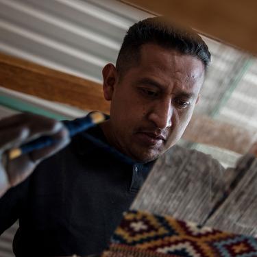 Seen through the warp of a loom, a man works on a woven textile.