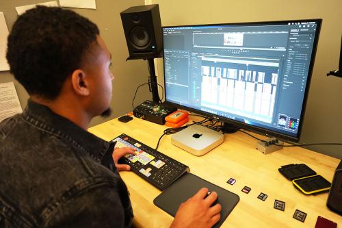 A young Black man sits at a computer, working with video-editing software. On the desk in front of him are five memory cards neatly lined up.