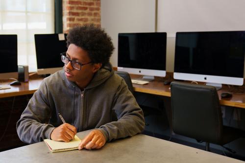 A young man writes notes in a notebook, with a row of Mac monitors behind him.